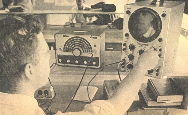 Charles is seated at an electronics workbench in high school where he is turning the control knobs on an oscilloscope. His face reflection can be seen in the glass of the oscilloscope.