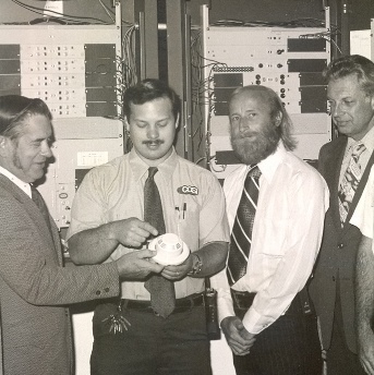 Charles is wearing a C O S I shirt and standing in from of the newly installed fire alarm system control panel along with other C O S I staff and contributors at a 1977 dedication ceremony.