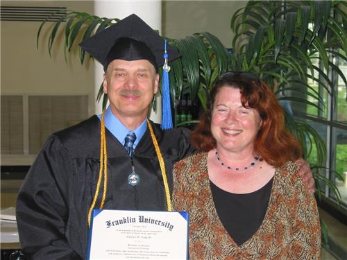 Charles is seen after the fall 2009 Franklin graduation. Charles is wearing a black cap and robe with blue tassel and gold honors cord while holding his diploma alongside his wife with red hair and wearing a brown and black blouse.