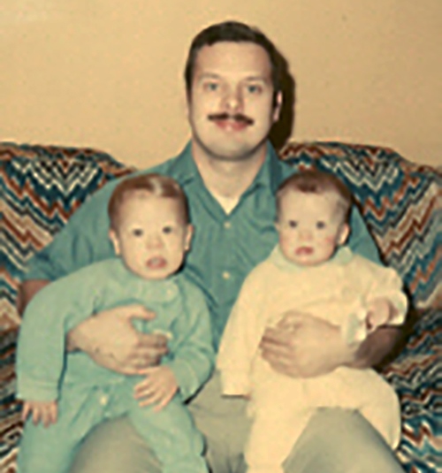 Charles is seated on a colorful couch in 1977 with a one year old Matthew on his right knee and a six month old Cathy on his left knee.