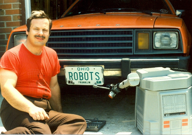 A Hero Junior robot is helping Charles install a personalized car license tag on his orange Ford Pinto in 1985. The license plate reads Robots.  